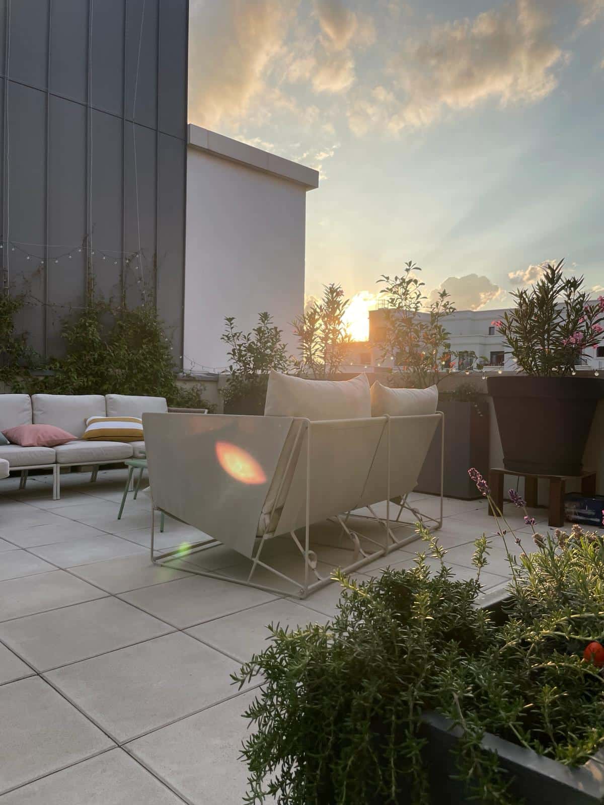 Grande terrasse de toit moderne au coucher du soleil, mobilier extérieur clair, plantes luxuriantes et lueur chaleureuse. Ambiance relaxante.