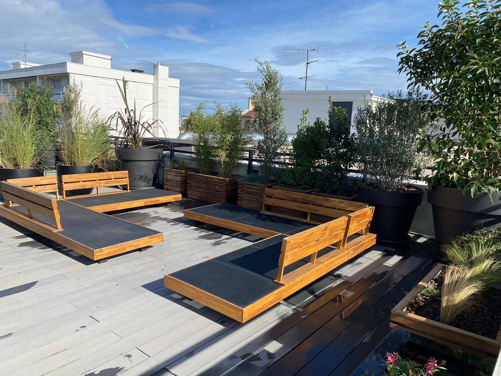 Terrasse de toit ensoleillée. Mobilier moderne en bois clair (transats, banquettes) sur decking gris. Grandes plantes en pots noirs et jardinières bois.