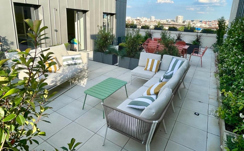 Terrasse de toit moderne avec canapés extérieurs beiges, coussins rayés jaunes et verts, table basse verte. Verdure luxuriante et vue dégagée sur la ville.