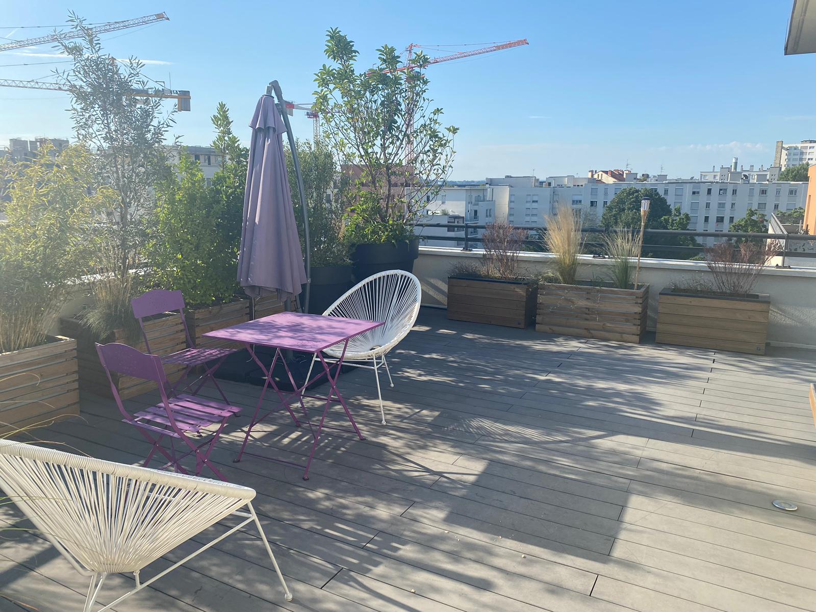 Terrasse ensoleillée sur toit avec mobilier de jardin violet et blanc, plantes en jardinières et vue sur la ville avec grues de construction.