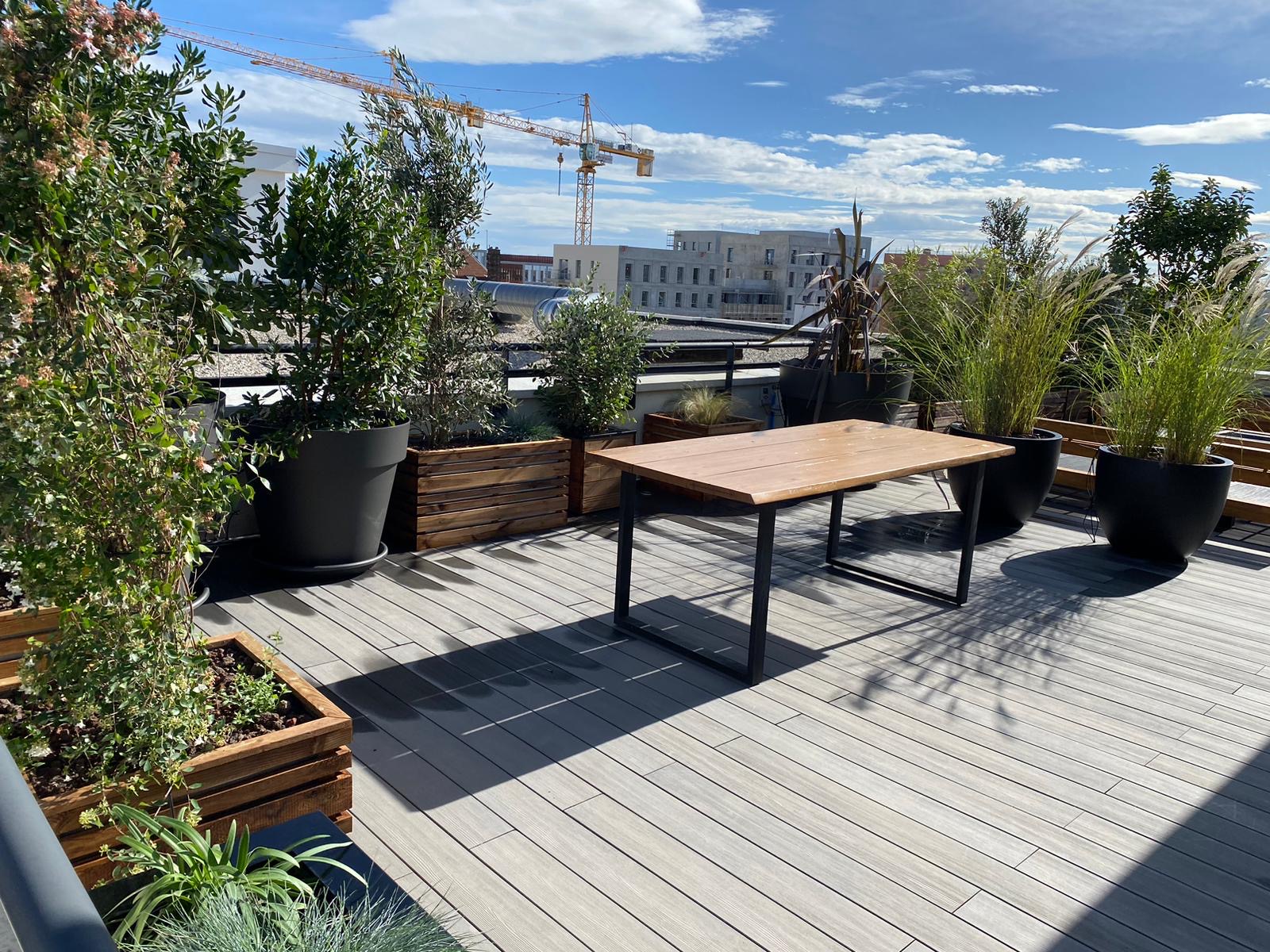Terrasse de toit moderne avec un plancher en bois clair, une table en bois et métal noir, et de grandes plantes vertes en pots. Ciel bleu, nuages et grues de chantier en arrière-plan.