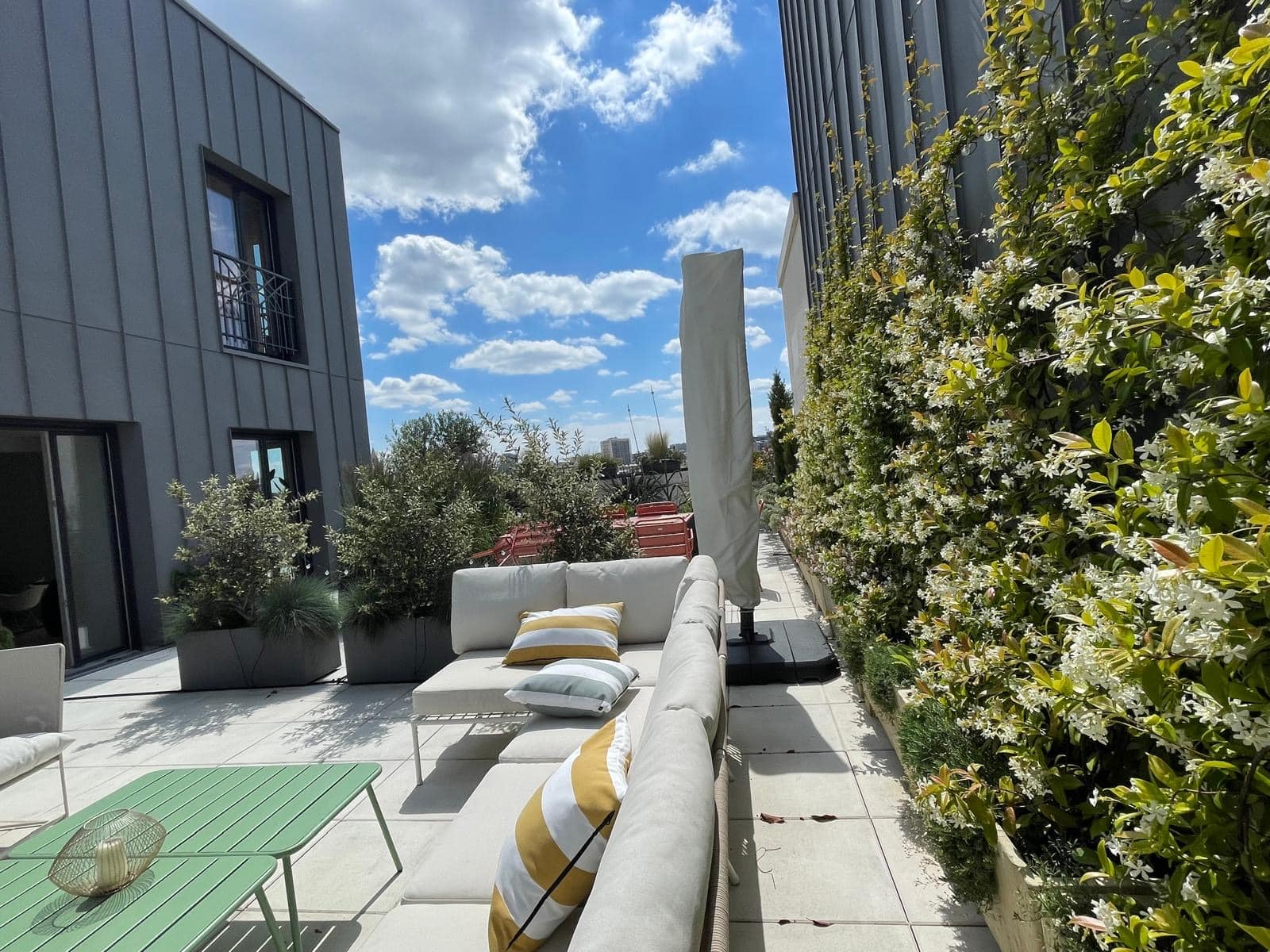 Terrasse de toit moderne et ensoleillée avec canapé beige, coussins rayés, table verte, murs végétalisés de jasmin en fleurs, ciel bleu.