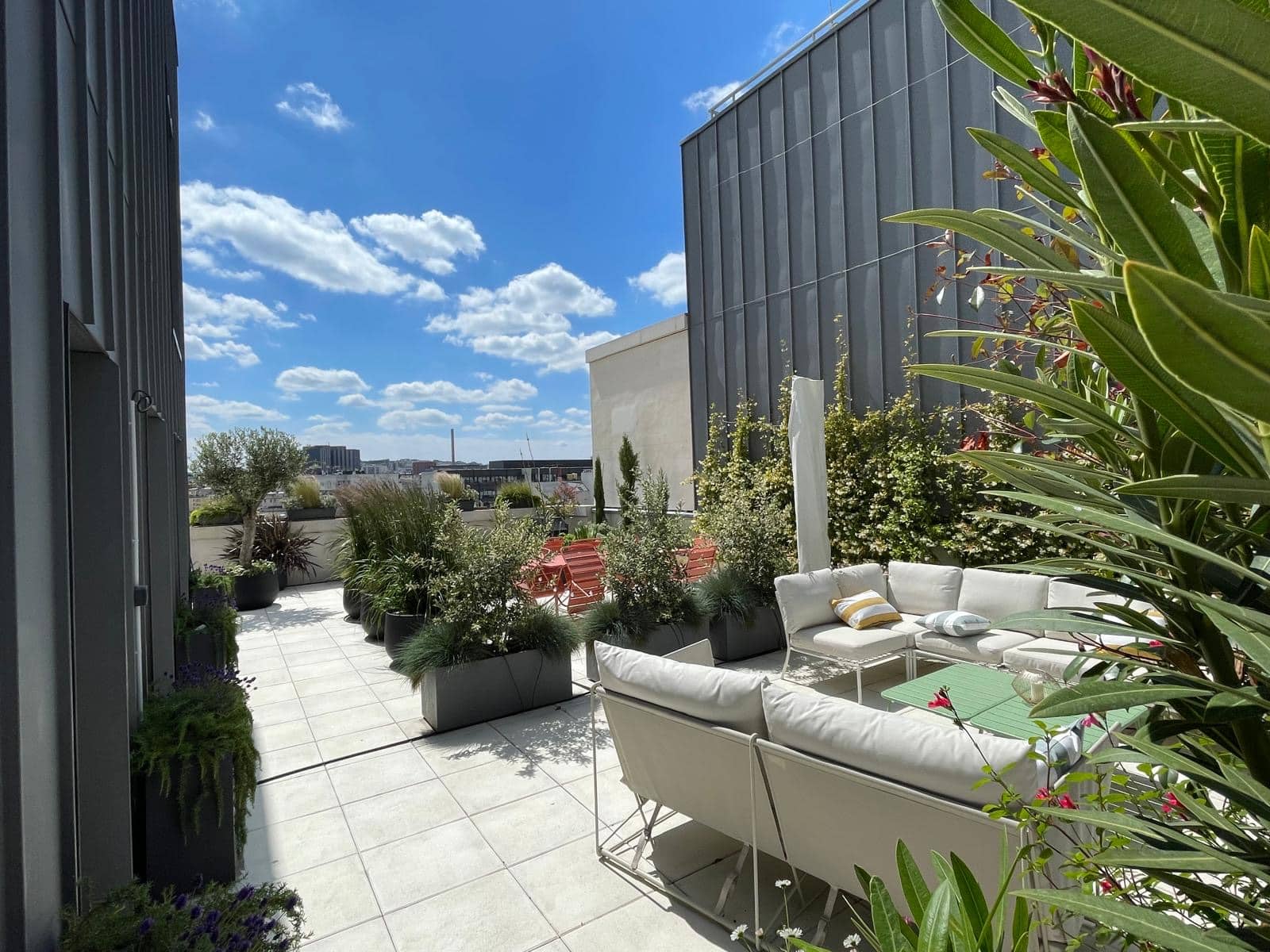 Luxueuse terrasse de toit urbaine avec canapés clairs, table basse verte et plantes verdoyantes. Ciel bleu, nuages, vue sur la ville moderne.