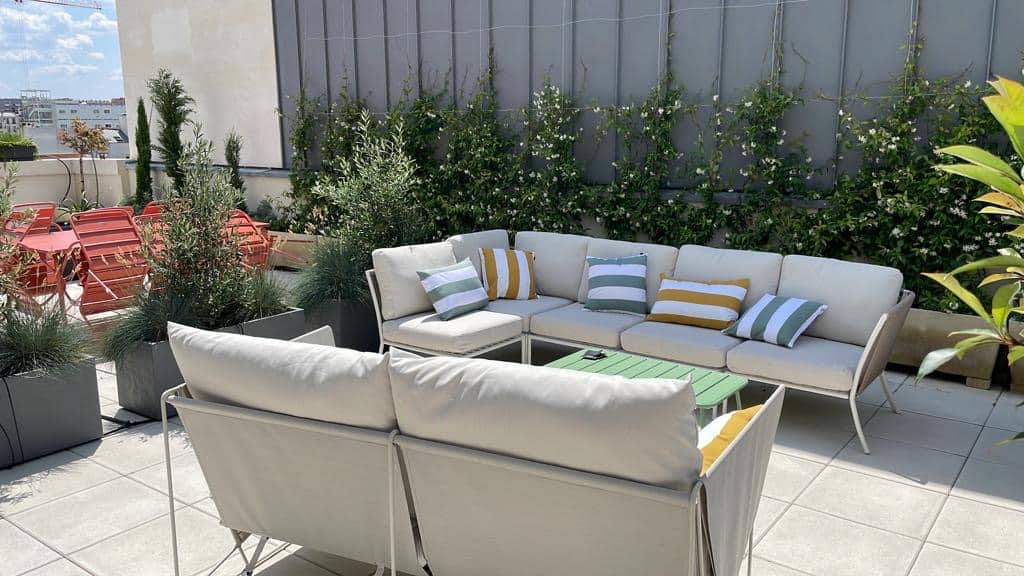 Terrasse de toit ensoleillée avec canapés extérieurs beiges, coussins rayés vert/jaune, table basse verte. Plantes et chaises rouges en arrière-plan.