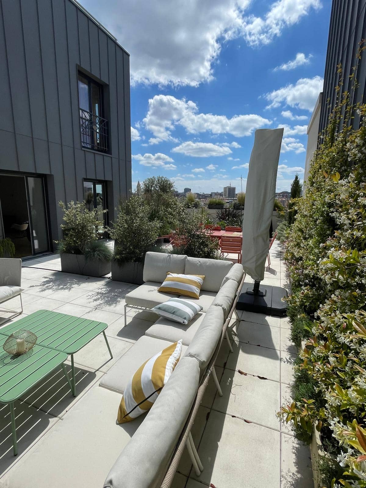 Terrasse sur toit moderne et ensoleillée avec canapé d'angle gris, table basse verte, plantes en bacs, et vue sur la ville sous un ciel bleu nuageux.