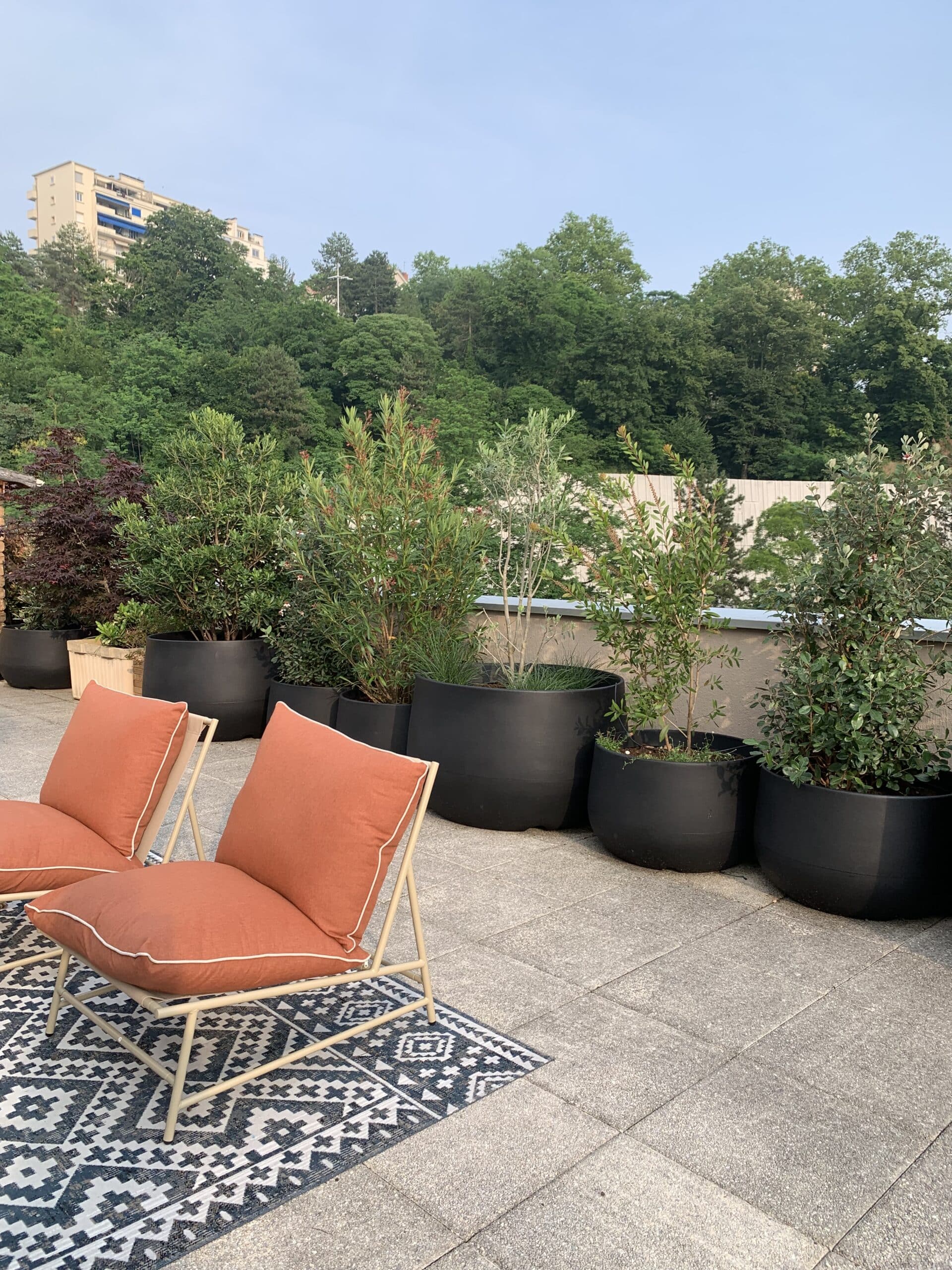 Terrasse moderne avec deux chaises lounge orange, tapis géométrique, plantes en pots noirs, et vue sur une colline boisée et un immeuble.