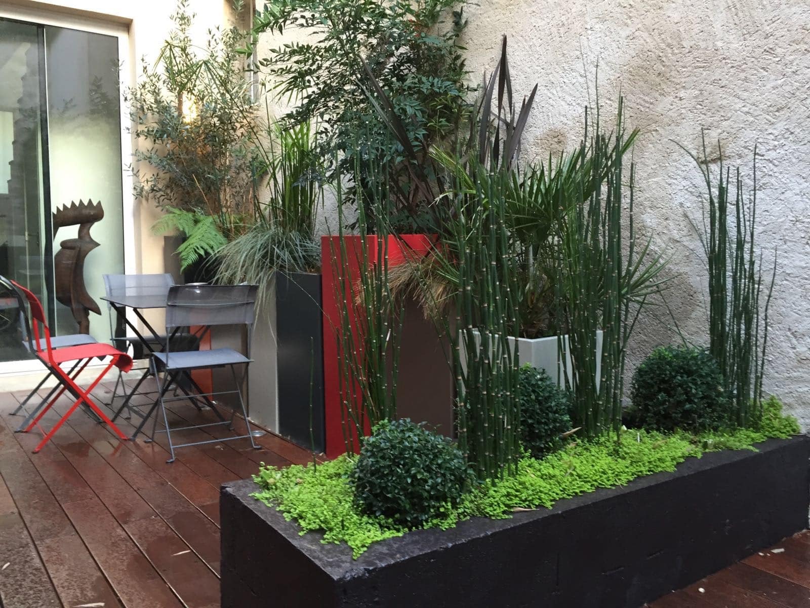 Jardin urbain contemporain. Longue jardinière noire de prêles et buis, devant des pots hauts noirs et rouges. Mobilier bistro sur deck bois.